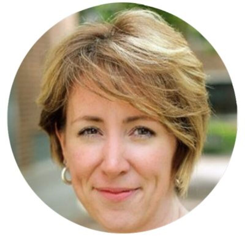 The image shows a woman with short, blonde hair and fair skin. She is wearing a light-colored top and hoop earrings. Her eyes are a light color, and she has a slight smile. The background is blurred, suggesting the photo was taken outdoors.